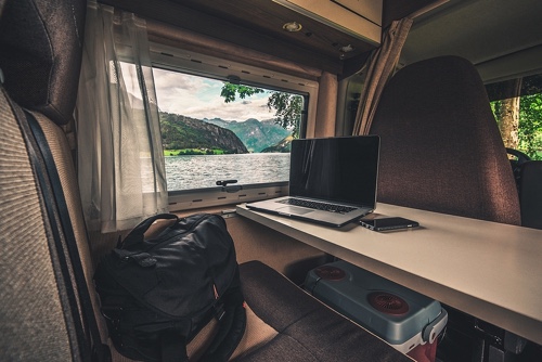 A laptop is shown sitting on a desk in an RV with a lake and mountains in the background