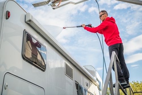 10 Ways To Avoid Expensive RV Repairs With Regular Maintenance with RV Masters in New Orleans/Kenner LA. Image of middle aged man standing on metal ladder in red hooded sweatshirt using power washing hose to wash off the top of the RV
