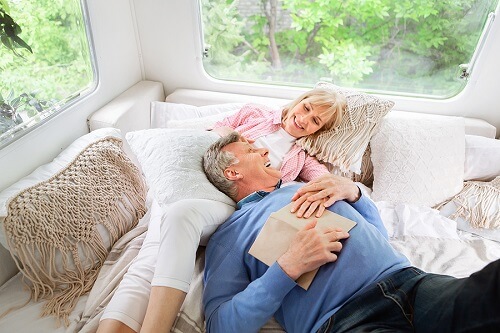 Why You Need A RV A/C Service And Repair Before Camping | RV Masters in Kenner, LA. Image of a happy senior couple relaxing in bed inside their cool RV during a camping vacation.