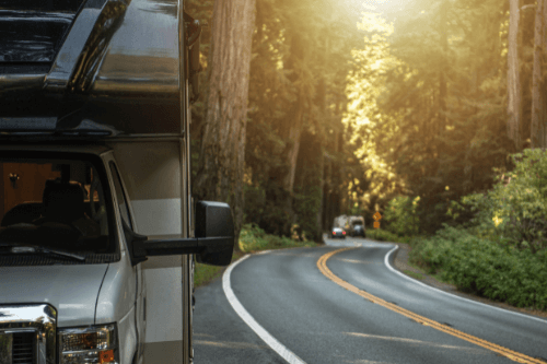 RV Preventative Maintenance Tips This Summer | RV Masters in Kenner LA. Image of a forest road with vehicles at a distance and a parked RV on the left.
